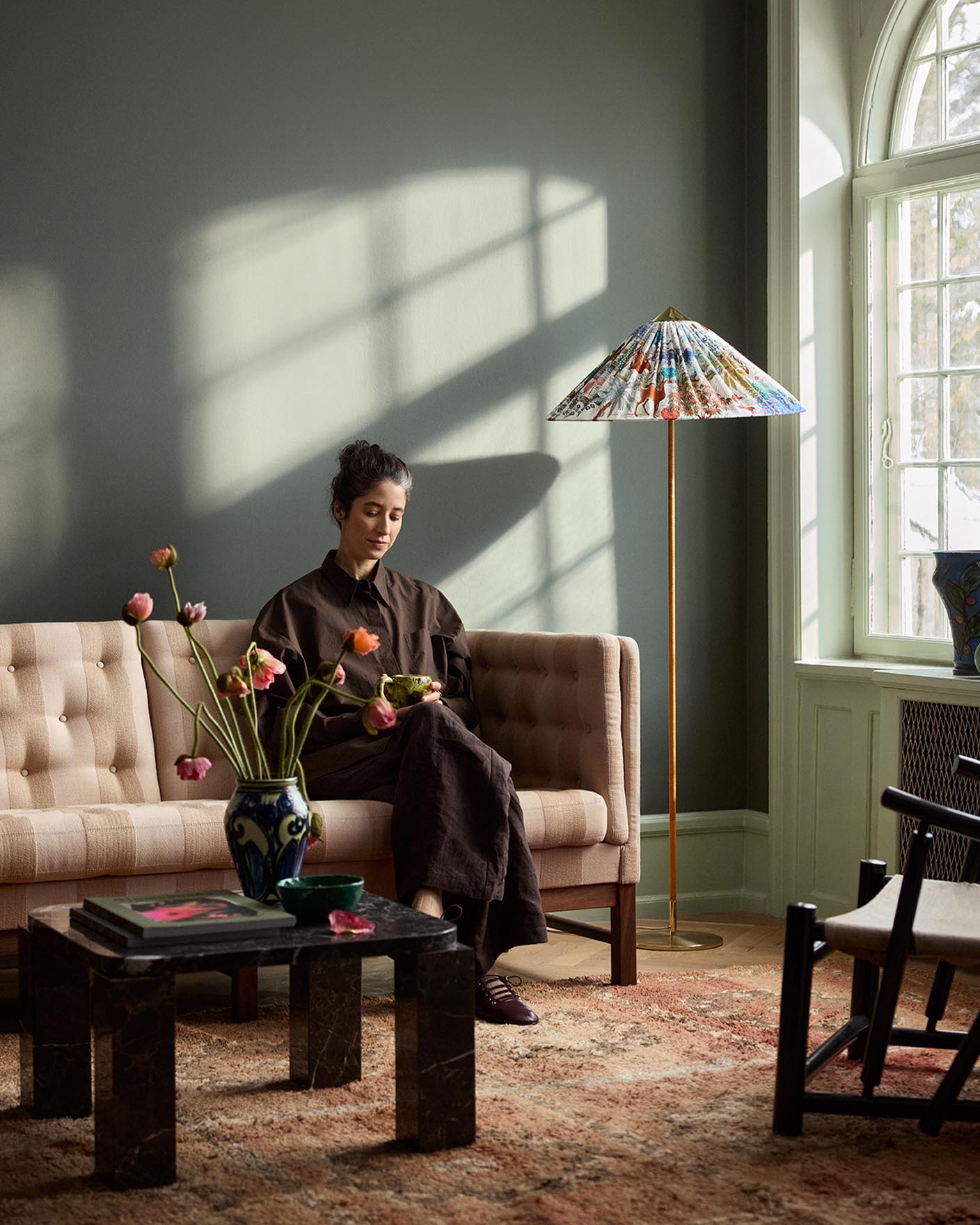 Woman sitting in a sofa with sunlight streaming through a window. The background wall is painted with the dark, muted green colour 7613 Northern Mystic from Jotun.