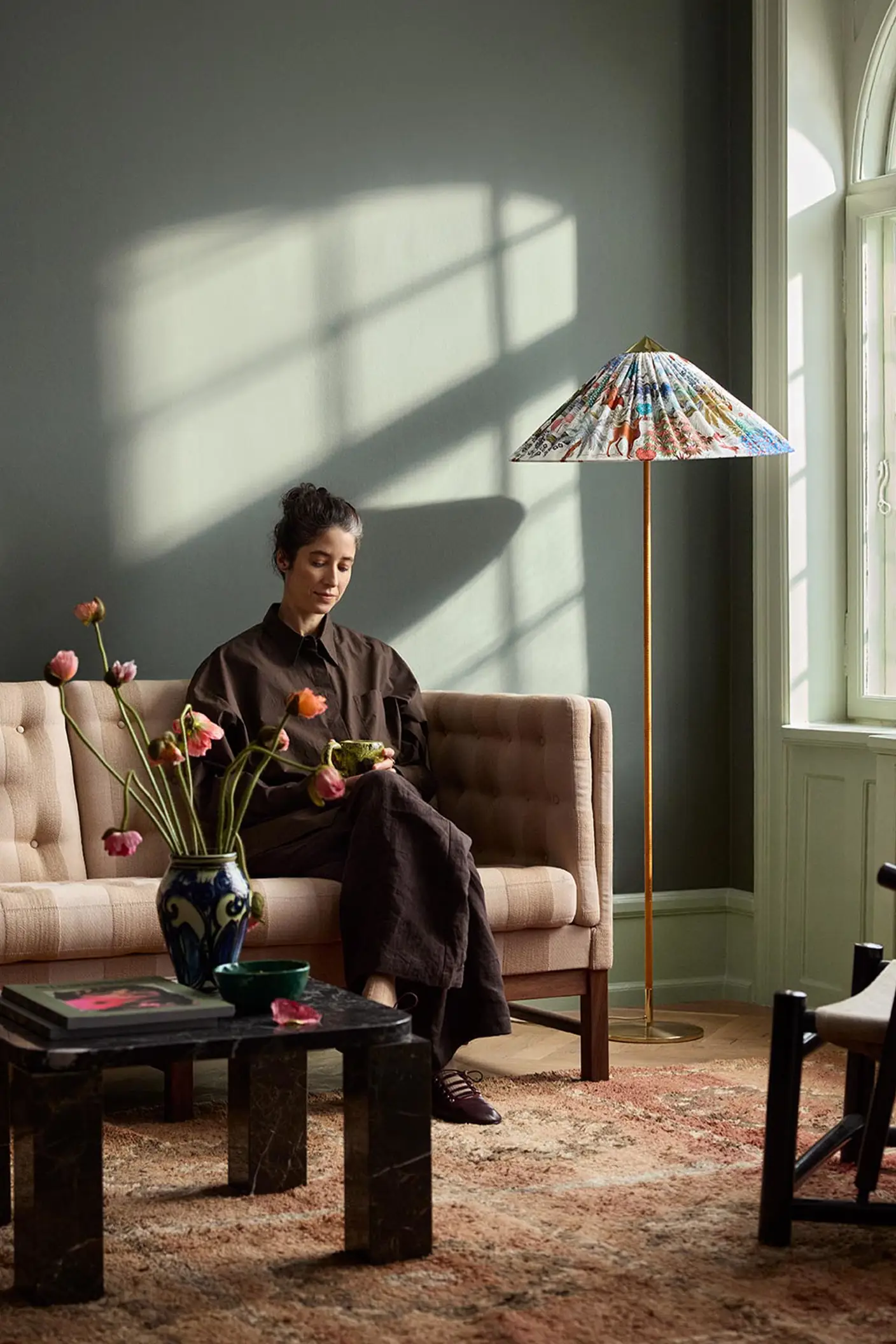Woman sitting in a sofa with sunlight streaming through a window. The background wall is painted with the dark, muted green colour 7613 Northern Mystic from Jotun.