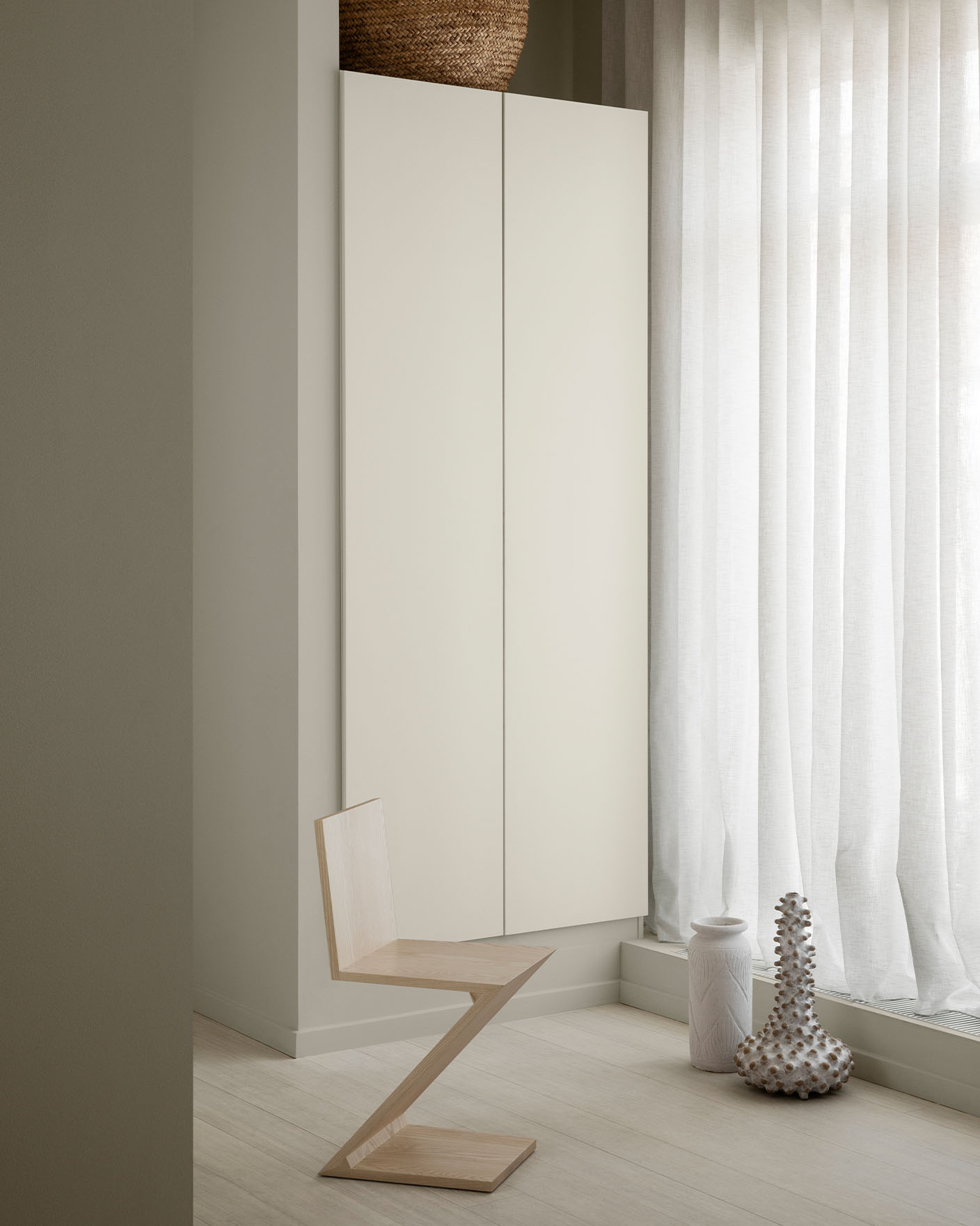Minimalist interior with a geometric wooden chair, a cabinetry painted in Jotun 1376 Mist, and sheer white curtains. Wall in front painted in 12180 Present, a muted beige from Jotun.