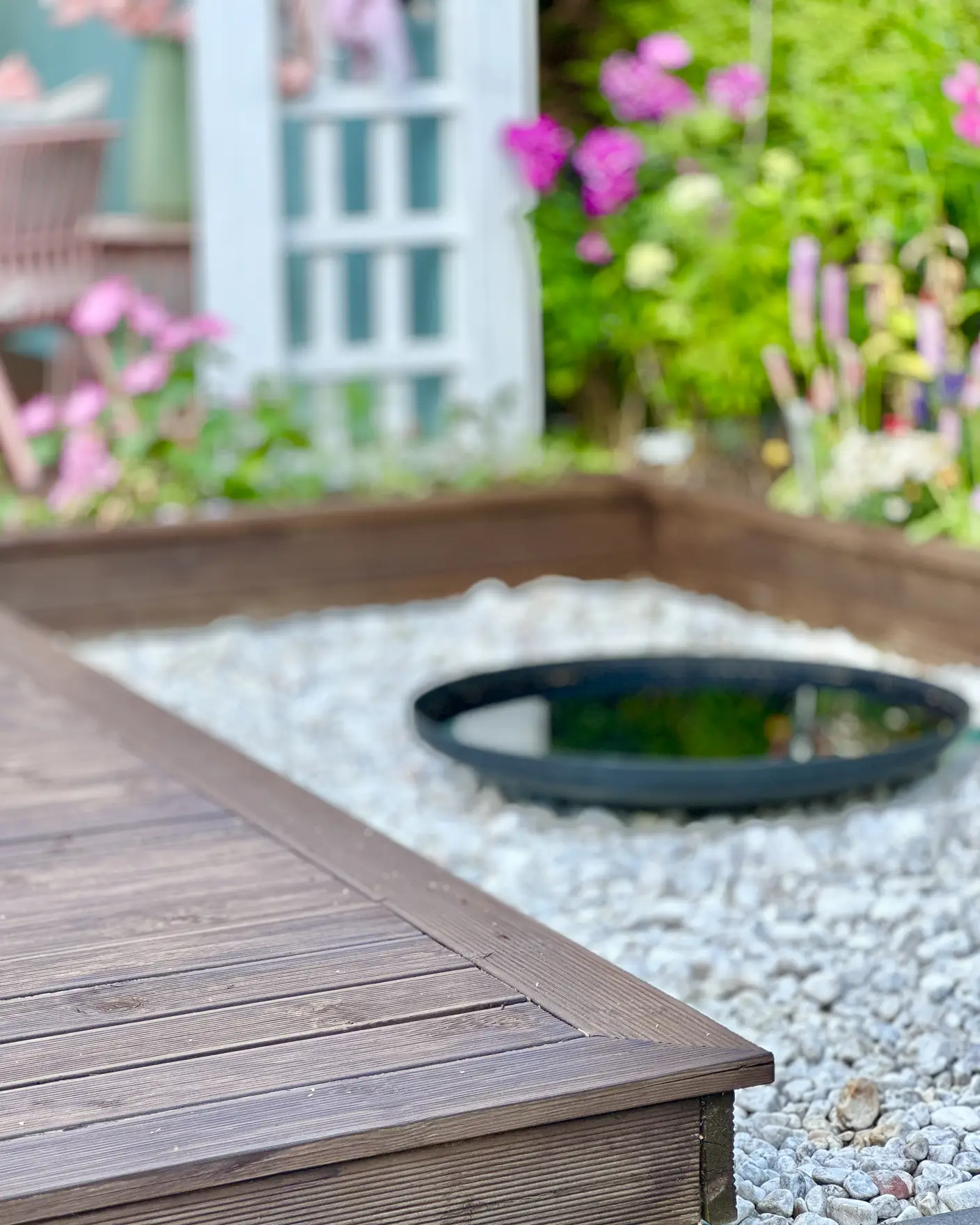 Detalj av brunbeiset terrasse i fargen TREBITT 90000 Terrassebrun mot hvite dekorstener og grønne vekster og rosa blomster.
