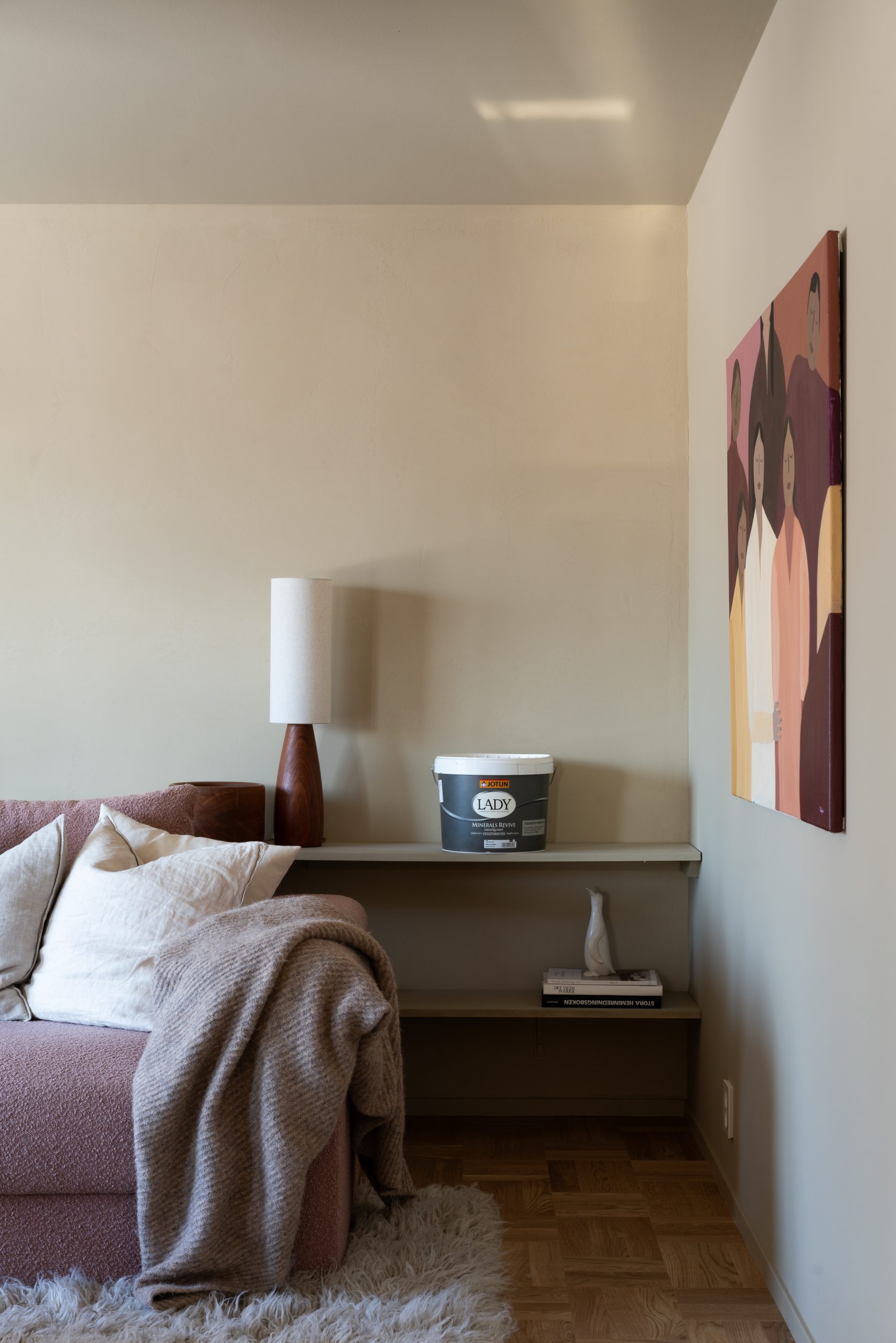 Moderne stueinteriør med beige vegger, en rosa sofa med hvite puter, en brun lampe, og et maleri i varme farger. Vegghylle med en bøtte LADY maling fra Jotun og pyntegjenstander