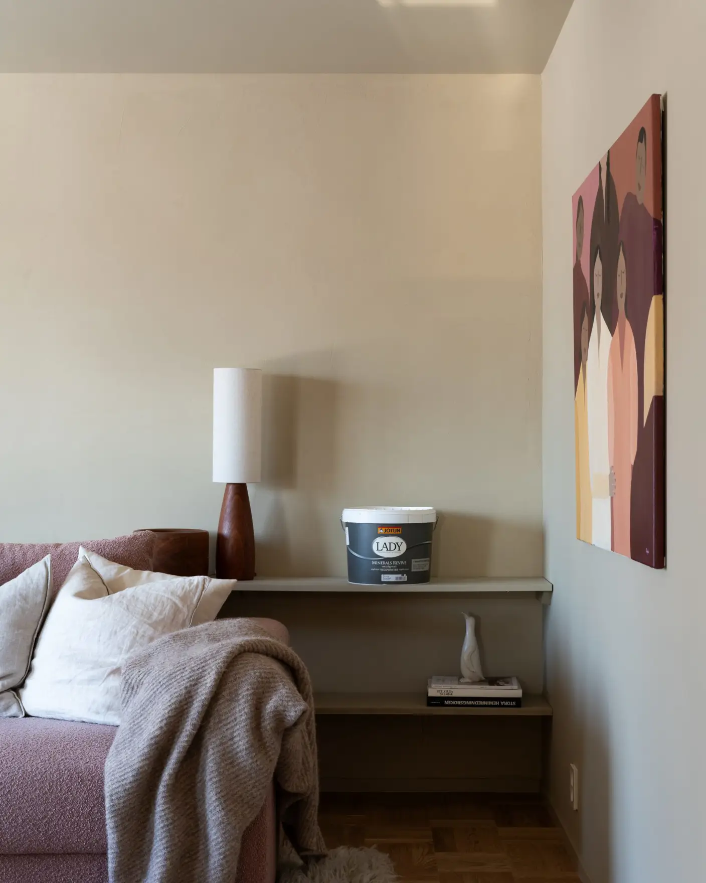 Moderne stueinteriør med beige vegger, en rosa sofa med hvite puter, en brun lampe, og et maleri i varme farger. Vegghylle med en bøtte LADY maling fra Jotun og pyntegjenstander