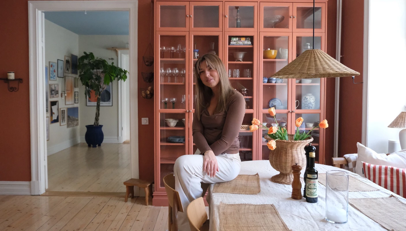 Kvinne sitter på spisebord med vitrineskap og vegg i bakgrunnen malt i den dempede korallfargen Jotun 20217 Muted Coral.