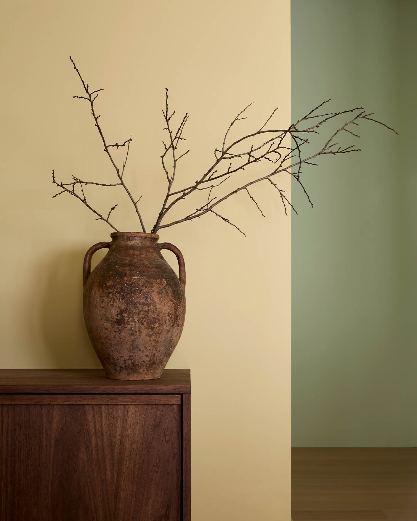 A rustic clay vase with bare branches sits on a wooden surface against a soft yellow wall in the colour Jotun Earthy Yellow in the front and a wall in Avocado green in the back.
