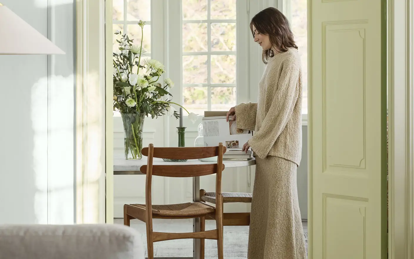 Woman in cozy beige clothing standing by a dining table with a vase of white flowers, in a sunlit room with large windows