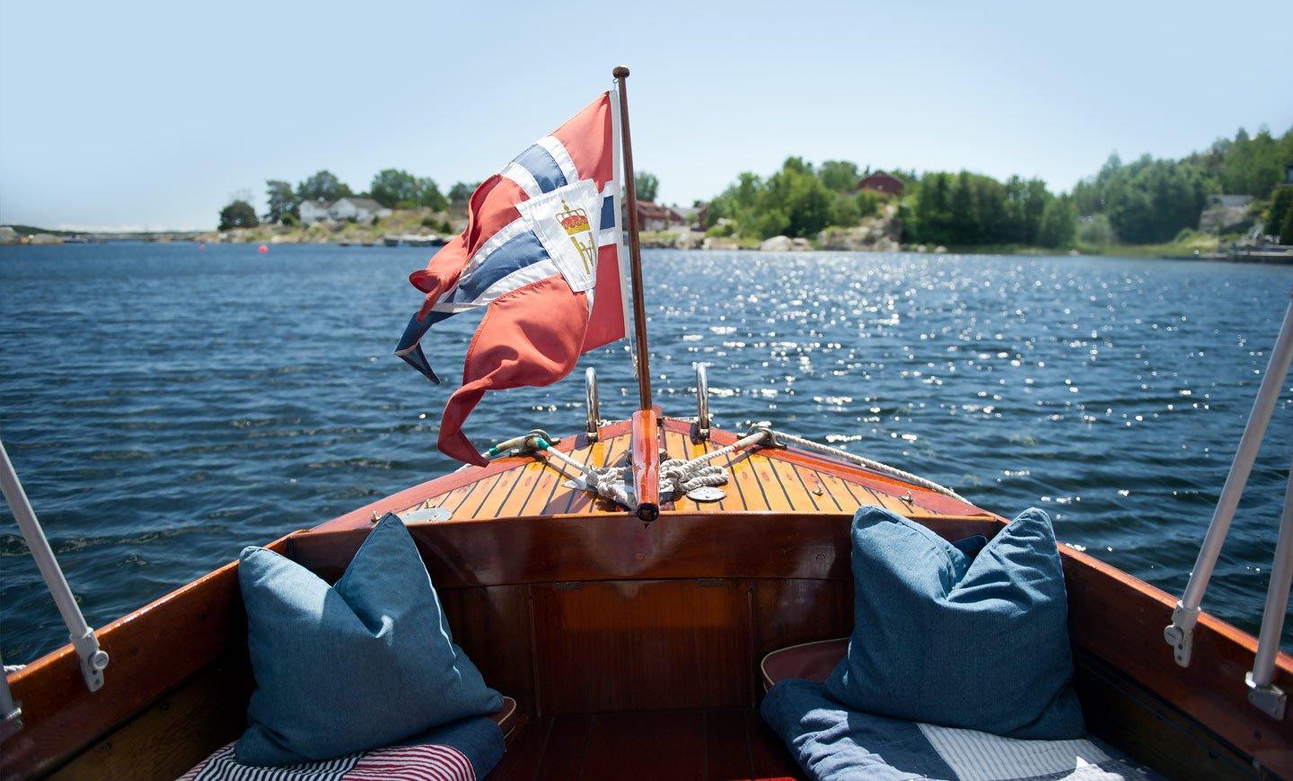 Norsk trebåt på sjøen med norsk flagg i brisen, omgitt av blått vann og grønne trær i bakgrunnen. To puter i båten