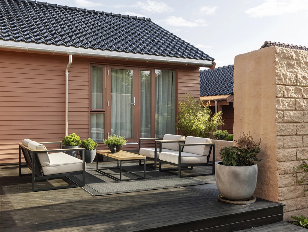 Moderne utendørs terrasse med sorte metallstoler og beige puter på en treplatting, ved siden av et hus med røde paneler malt i Jotun 20118 Amber Red og store vinduer, omgitt av potteplanter og en steinmur