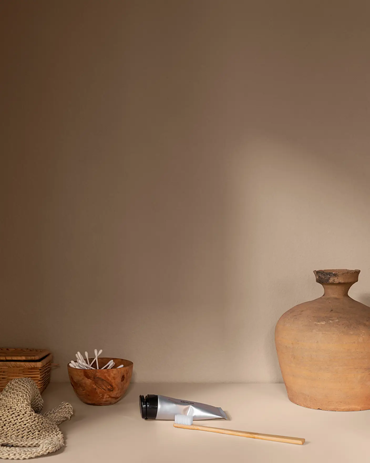 Keramisk vase ved siden av en flettet kurv, en bolle med bomullspinner, tannkremtube og bambustannbørste på en gyllen rosa benk malt i fargen Devine fra Jotun