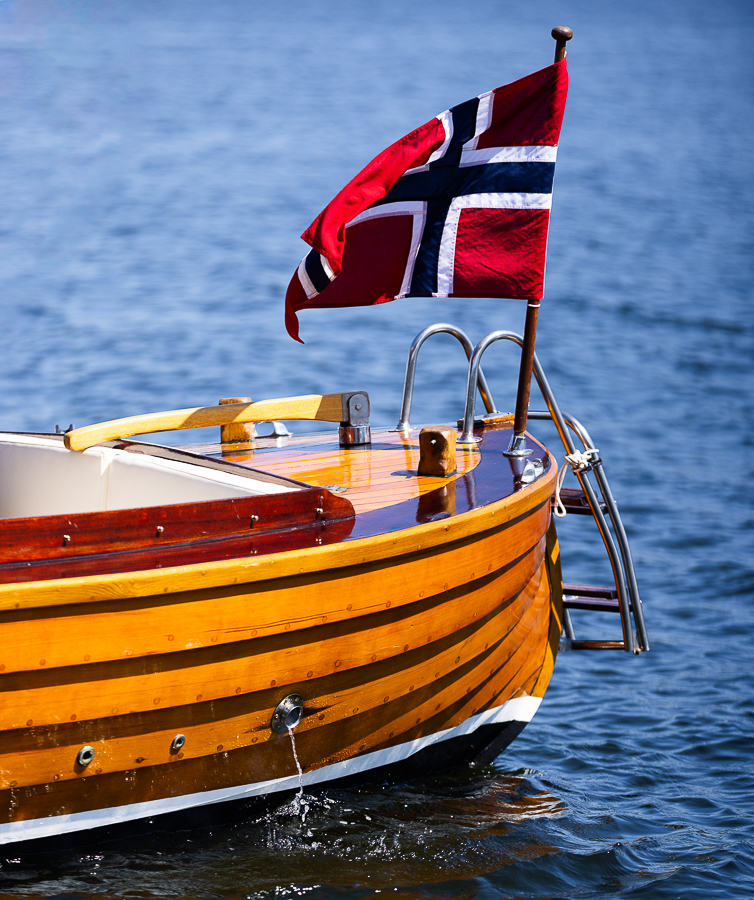Akterdekk på trebåt med norsk flagg som ligger på rolig vann i sommersol.