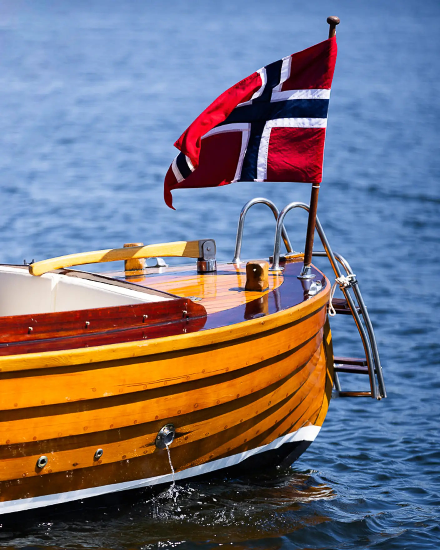 Akterdekk på trebåt med norsk flagg som ligger på rolig vann i sommersol.