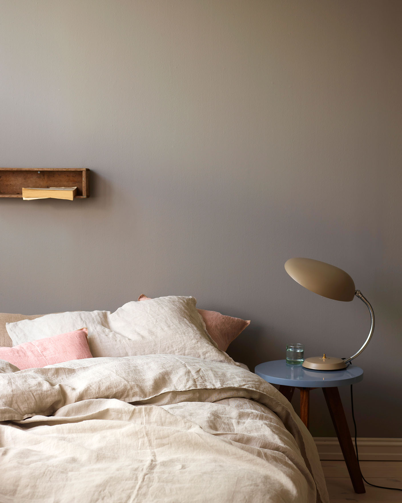 Neutral-toned bed with soft linen bedding and pale pink accent pillows, beside a  side table holding a retro lamp, against a golden grey wall painted with Jotun 1877 Pebblestone.