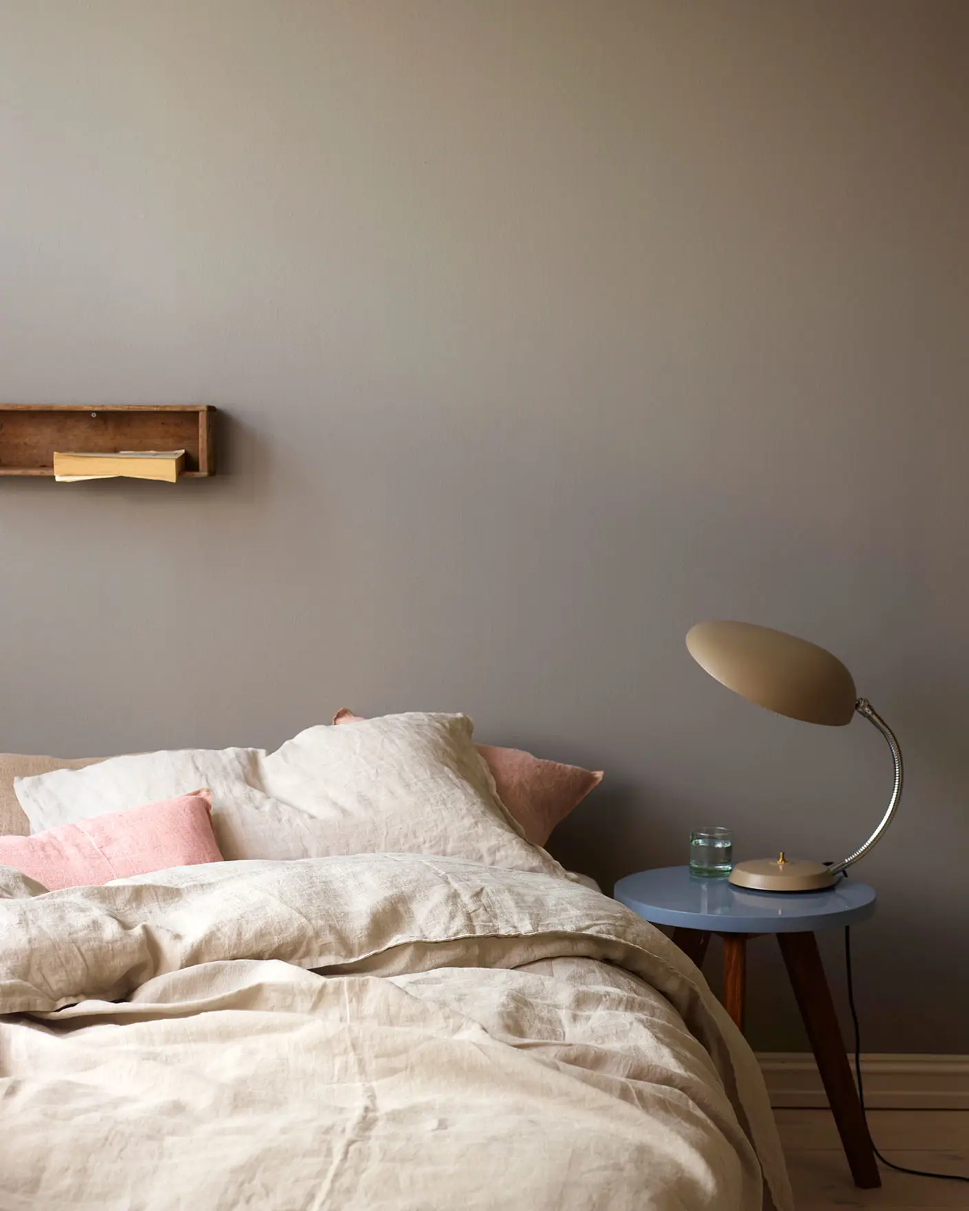 Neutral-toned bed with soft linen bedding and pale pink accent pillows, beside a side table holding a retro lamp, against a golden grey wall painted with Jotun 1877 Pebblestone.