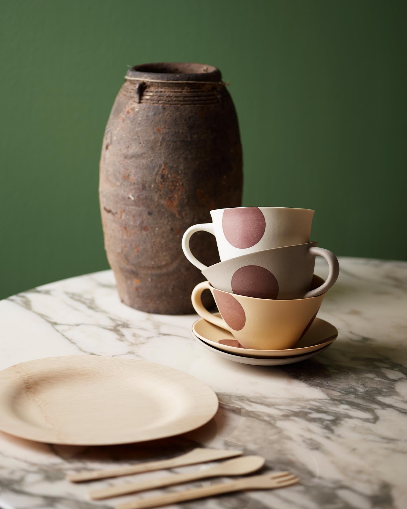 Stablede kopper og rustikt keramikk på et marmorbord, mot en gulgrønn vegg malt med Jotun 0125 Palmetto for en koselig, naturlig følelse.