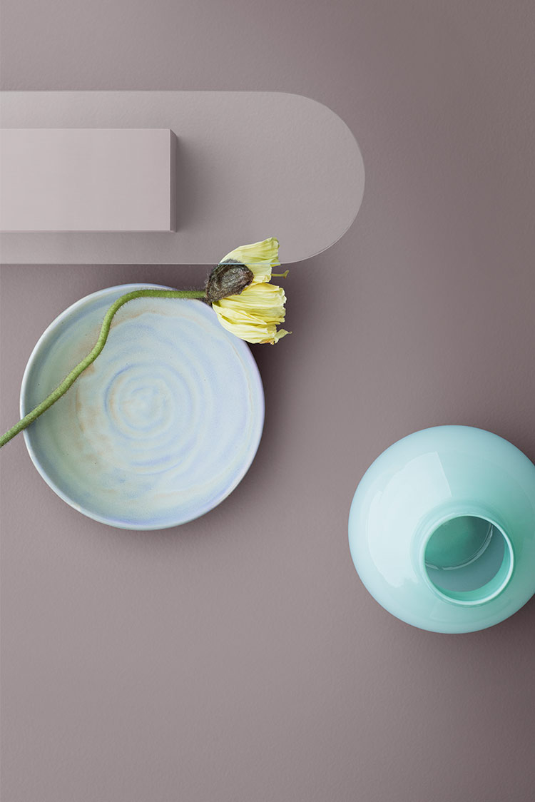 Minimalistisk stillebenkomposisjon med en blå keramisk bolle, en lysegrønn vase og en gul blomst på en dempet brun bakgrunn