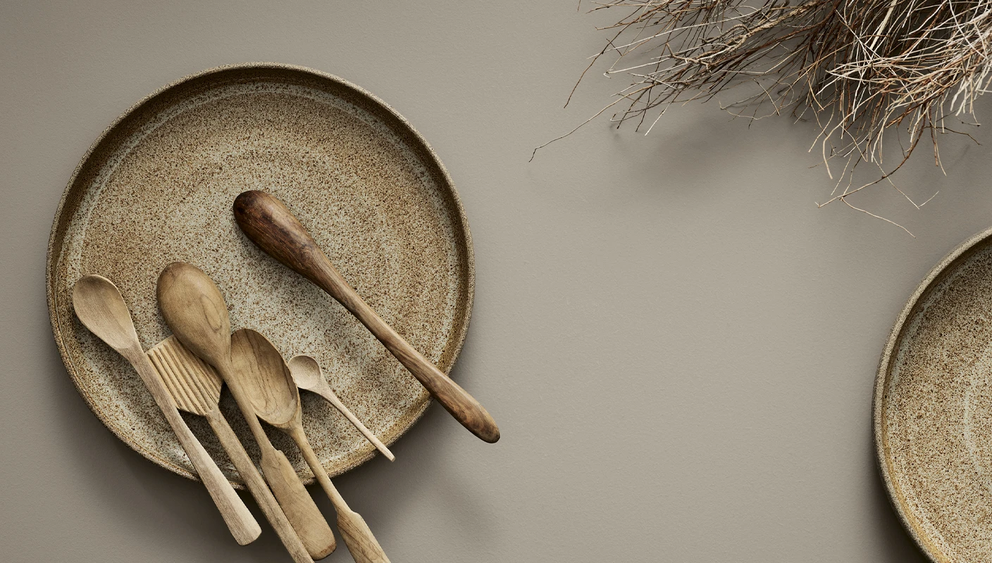 Flatlay med keramikktallerken i beige med treskjeer, liggende på en matt overflate malt i brunbeige Jotun Shade.