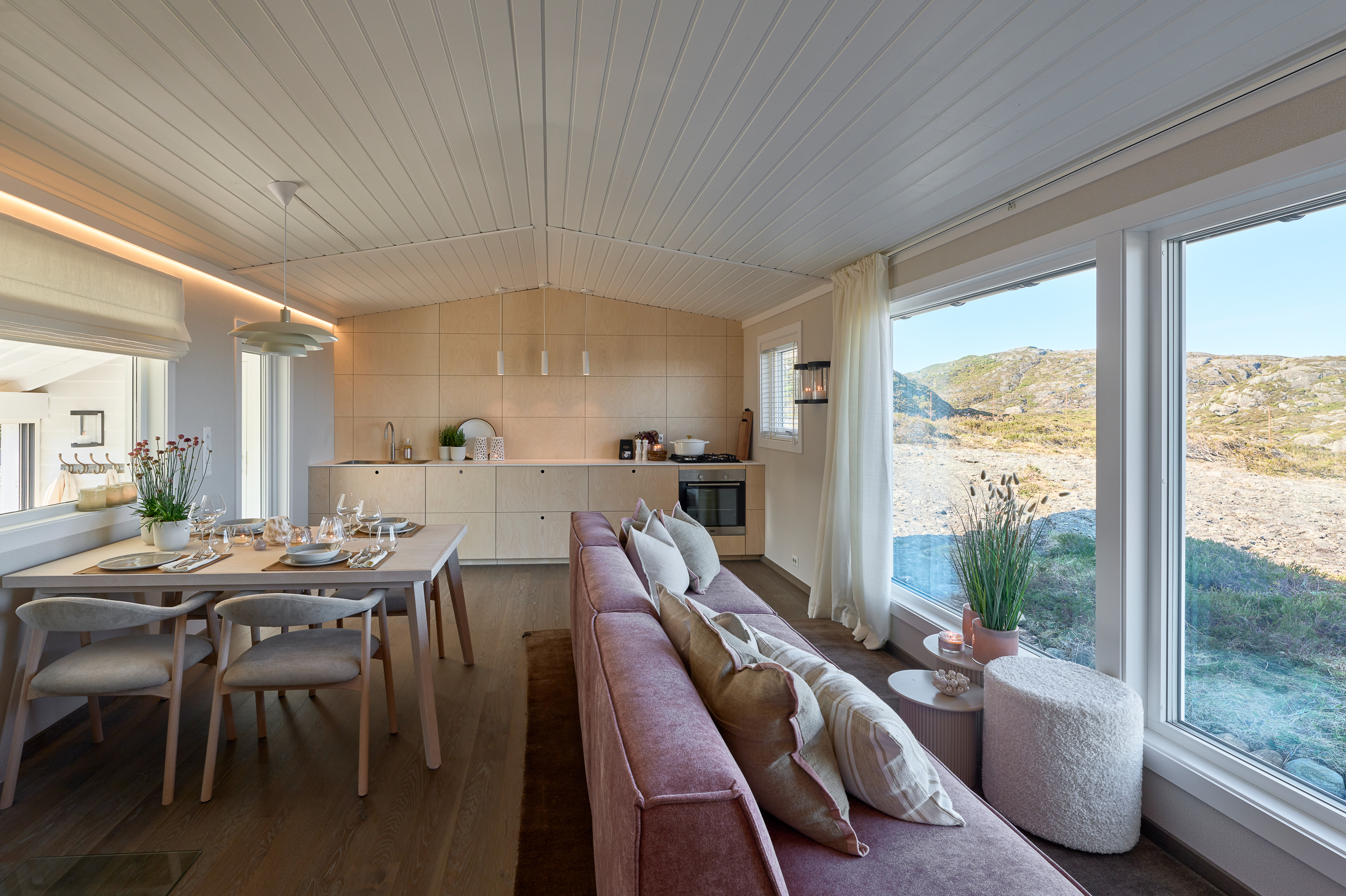 Moderne interiør i stue med store vinduer som gir utsikt til naturen, hvitt tak, åpen kjøkkenløsning og spisebord med fire stoler