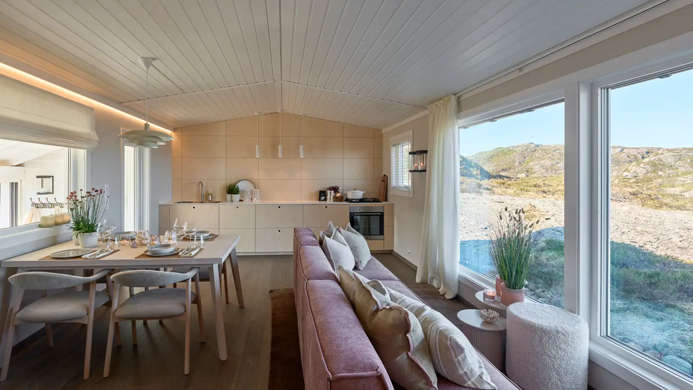 Moderne interiør i stue med store vinduer som gir utsikt til naturen, hvitt tak, åpen kjøkkenløsning og spisebord med fire stoler