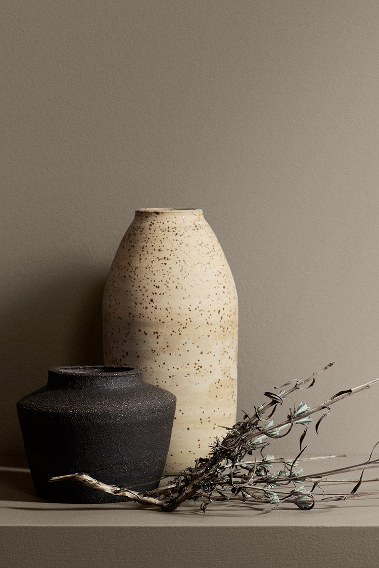 To keramiske vaser, den ene flekkete beige og den andre matt svart, plassert på en beige overflate med tørkede grener, som viser minimalistisk og rustikk dekor, mot en greige vegg malt i Jotun 11130 Shade