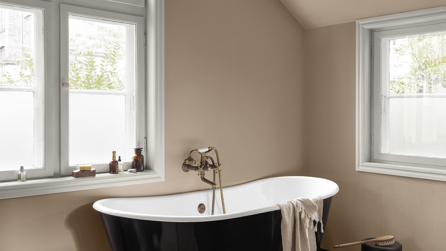 Badekar i elegant baderom med beige vegger malt med Jotun LADY Aqua våtromsmaling i fargen Devine, plassert under et stort vindu. Naturlig lys skaper en avslappende atmosfære, mens flasker med toalettartikler står på vinduskarmer rundt badekaret