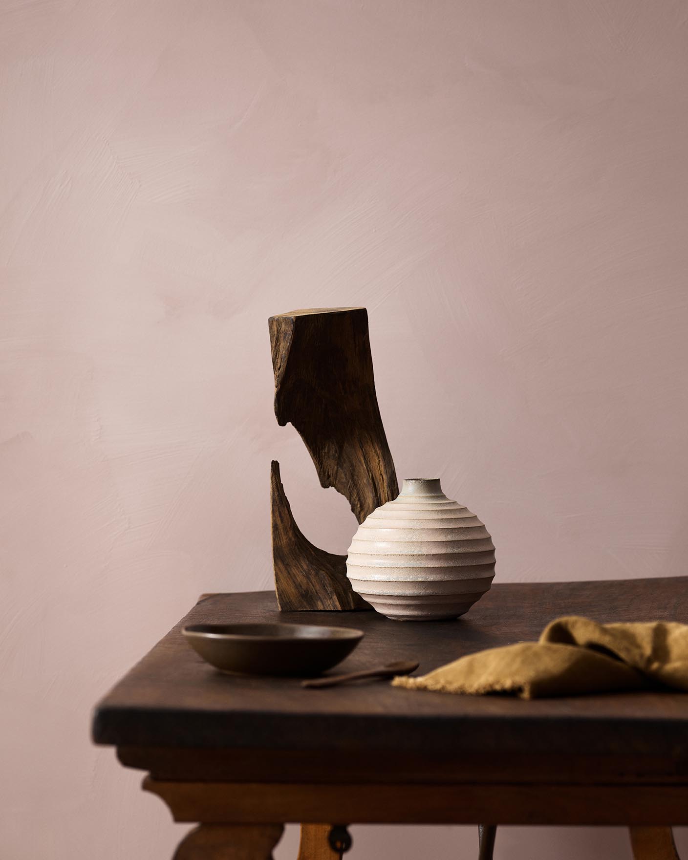 Wooden table with a ceramic vase, sculpted wood, and a dark bowl against a muted pink background painted with LADY Design Touch of Suede in the colour 20054 Silky Pink from Jotun.