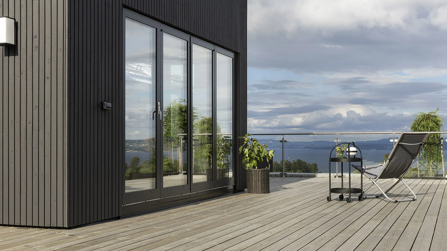 Moderne utendørs terrasse med tregulv beiset med TREBITT Terrassebeis i fargen 90029 Naturlig Sølvgrå, glassrekkverk og lenestol med utsikt over et naturskjønt landskap under en skyet himmel