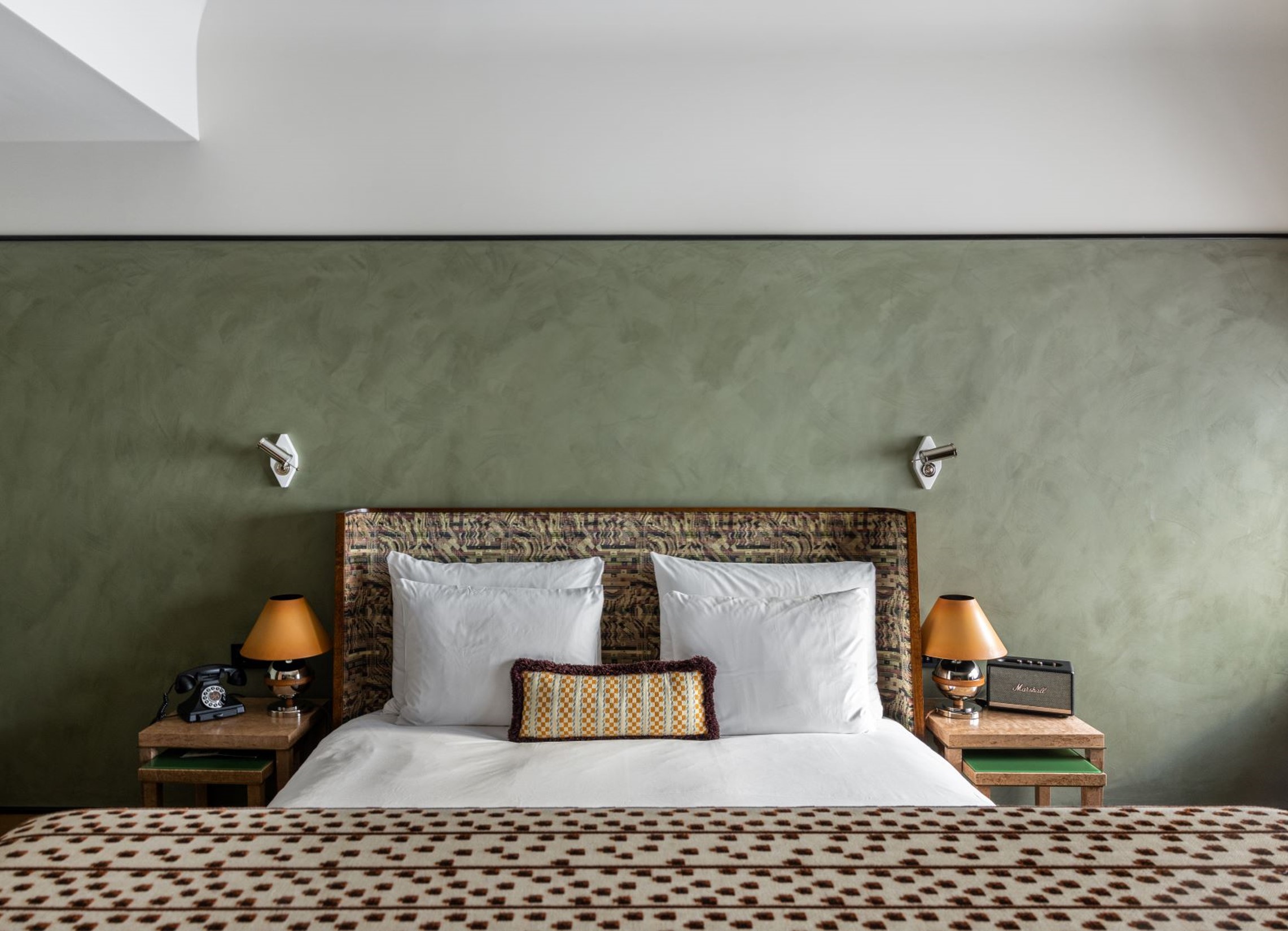 Koselig hotellrominteriør med en rustikk sengegavl, hvitt sengetøy, dekorative puter, grønn kalkmalt vegg med LADY Minerals kalkmaling i fargen , vintage telefon, bordlamper og radio på nattbord