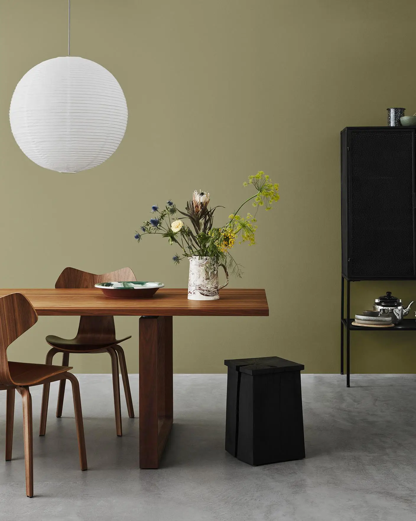 Et minimalistisk spisestue med tremøbler mot en olivengrønn vegg, malt med Jotun 8546 Local Green, og betonggulv.