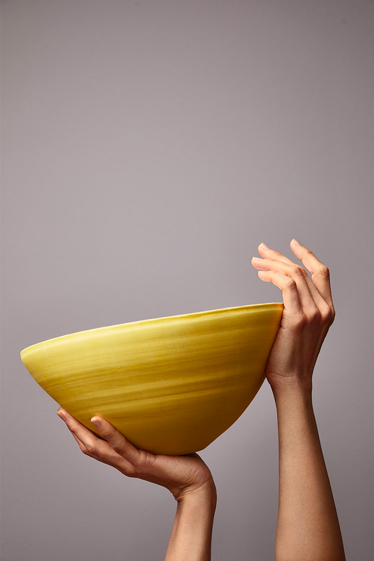Hender som holder en stor, gul keramikkbolle mot en grålilla bakgrunn malt i Jotun 3377 Slate Lavender
