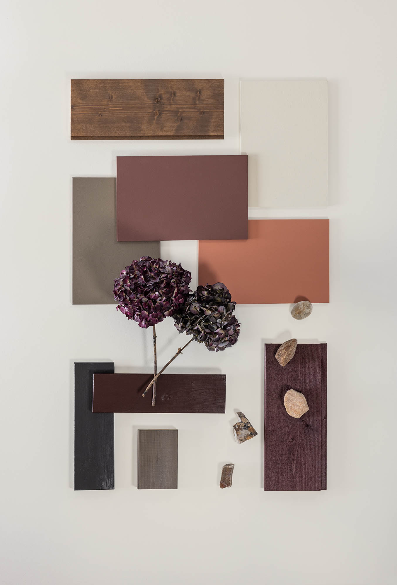 Fargeprøver i treverk og maling i brune, beige og rustrøde nyanser arrangert sammen, med lilla blomster og små steiner på en lys bakgrunn