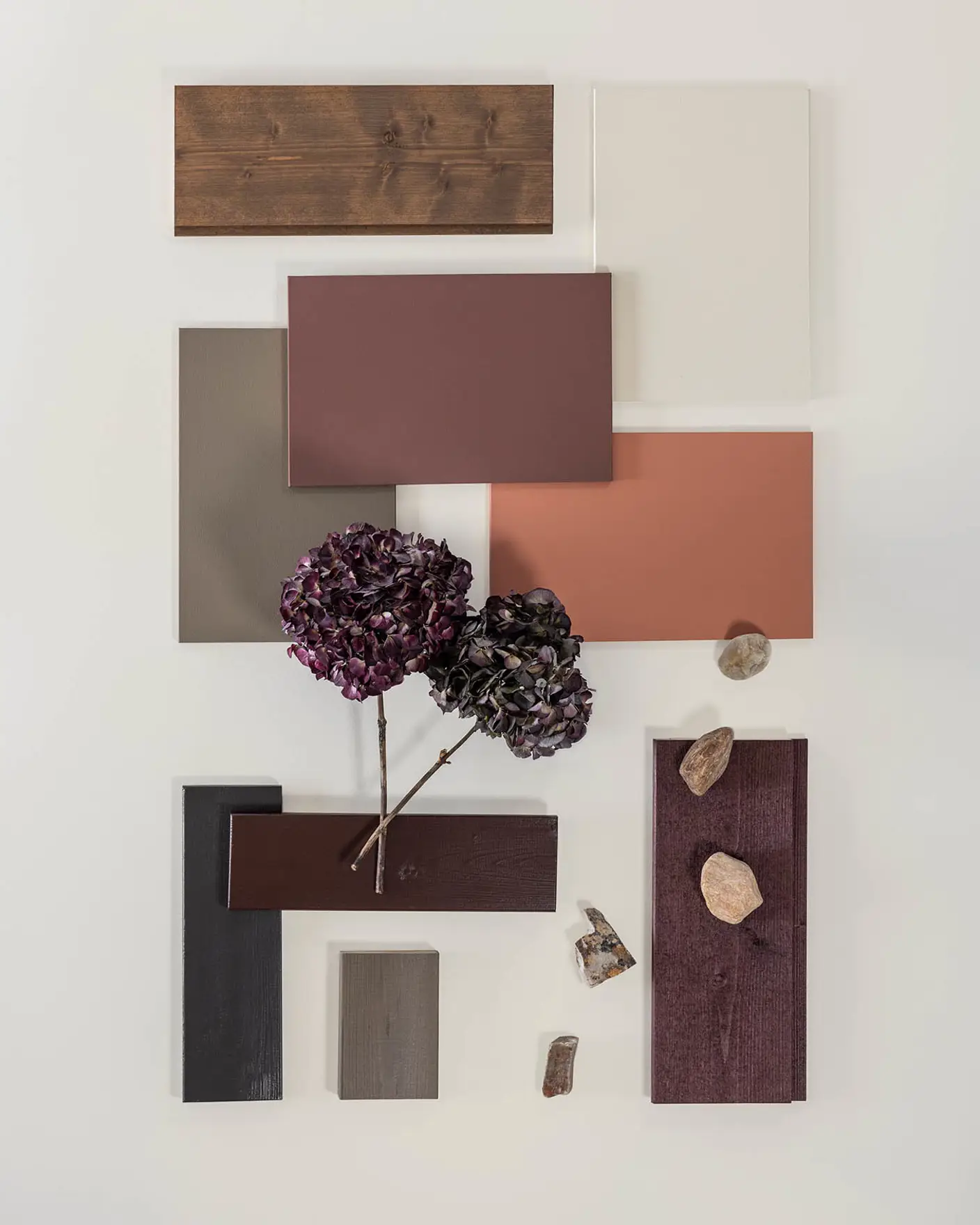 Fargeprøver i treverk og maling i brune, beige og rustrøde nyanser arrangert sammen, med lilla blomster og små steiner på en lys bakgrunn