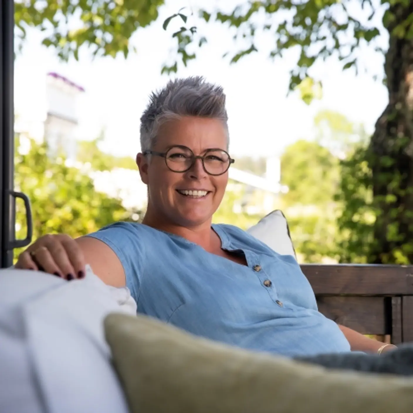 Line M. Bergan, fargerådgiver i Jotun. Kvinne med kort grått hår og briller smiler mens hun slapper av i en sofa utendørs, omgitt av grønt løv i solfylt vær