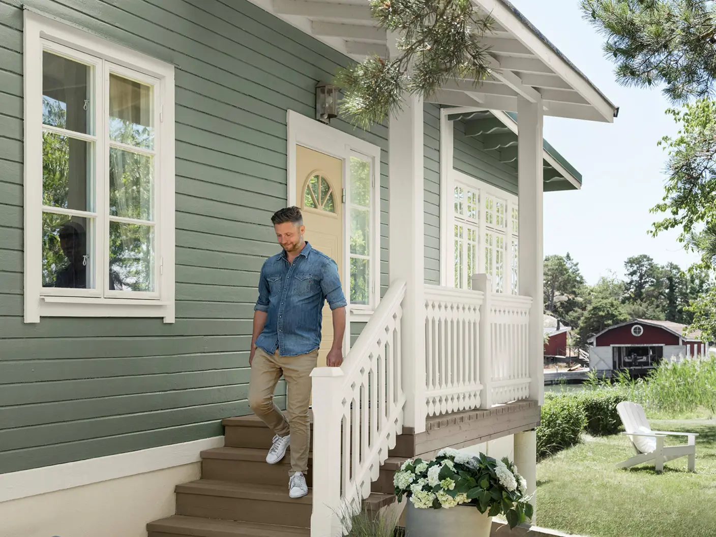 Man klädd i jeansskjorta som går nerför trappan vid grönt hus målad i Jotun 7653 Stockholmsgrön med vit veranda, omgiven av grönskande trädgård och utomhusstolar