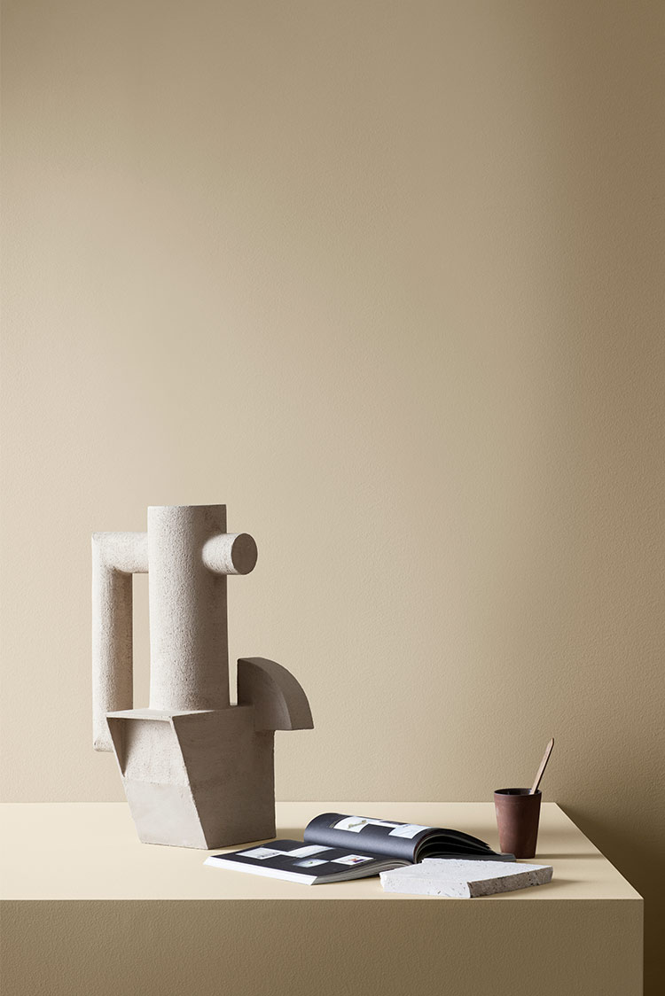 Abstrakt beige skulptur på minimalistisk bord ved siden av en åpen bok, steinplate og liten mørk kopp med blyant, mot en nøytral bakgrunn malt i beige Jotun 1303 Observe