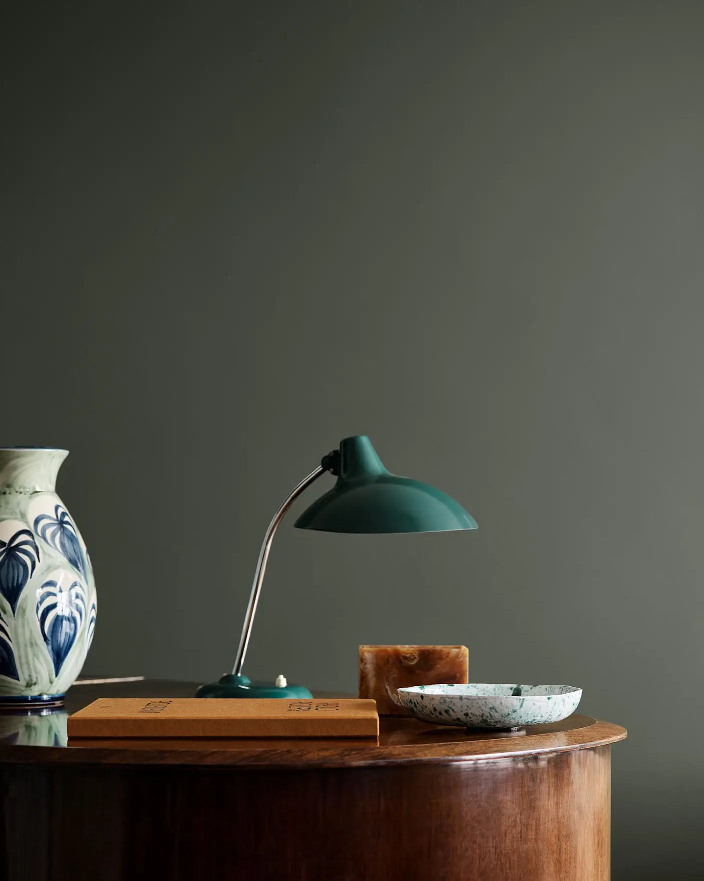 Wooden table with ceramic vase, lamp, and decor against a dark, muted green wall painted in the colour Jotun 7613 Northern Mystic.
