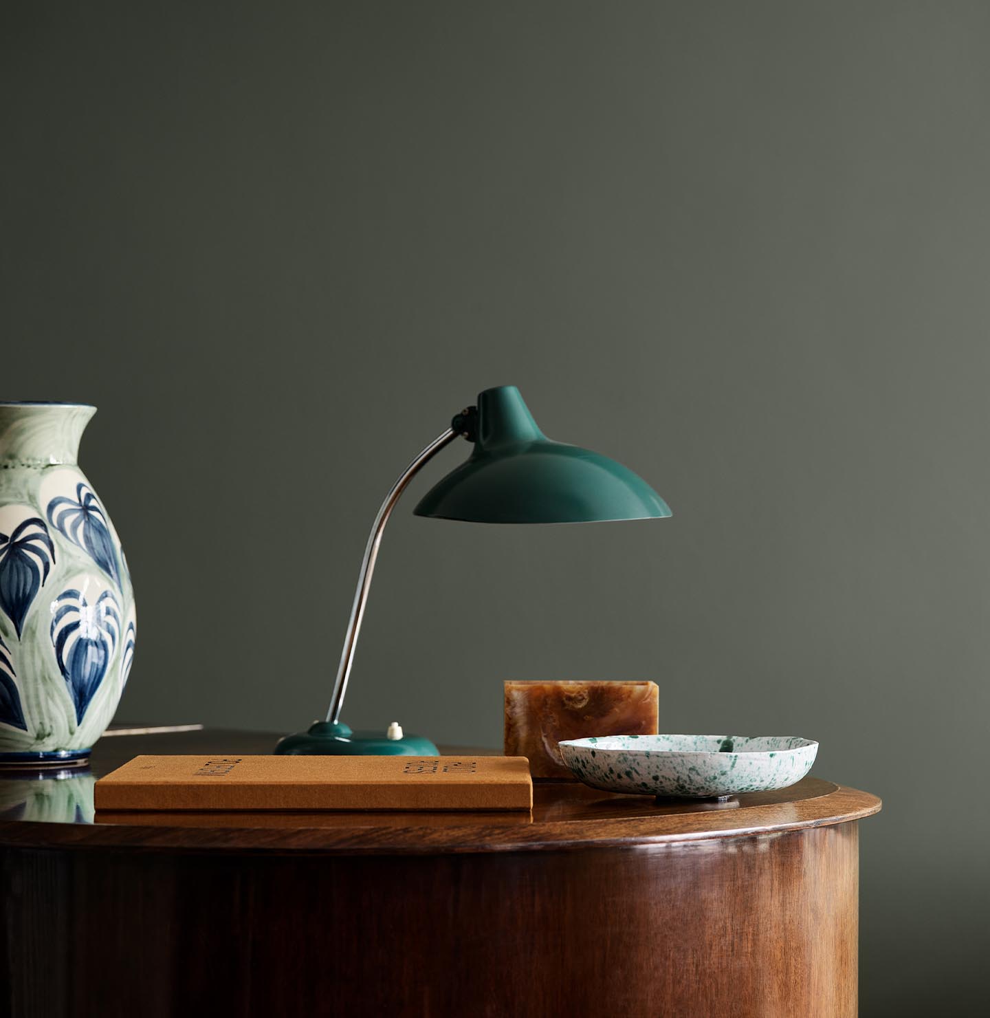 Wooden table with ceramic vase, lamp, and decor against a dark, muted green wall painted in the colour Jotun 7613 Northern Mystic.