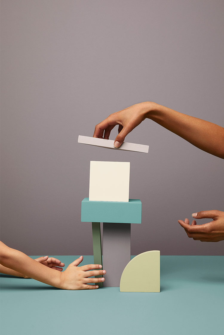 To hender bygger et tårn med geometriske treklosser på en turkis overflate malt i Jotun 6084 Sea Emerald mot en grålilla bakgrunn malt i Jotun 3377 Slate Lavender