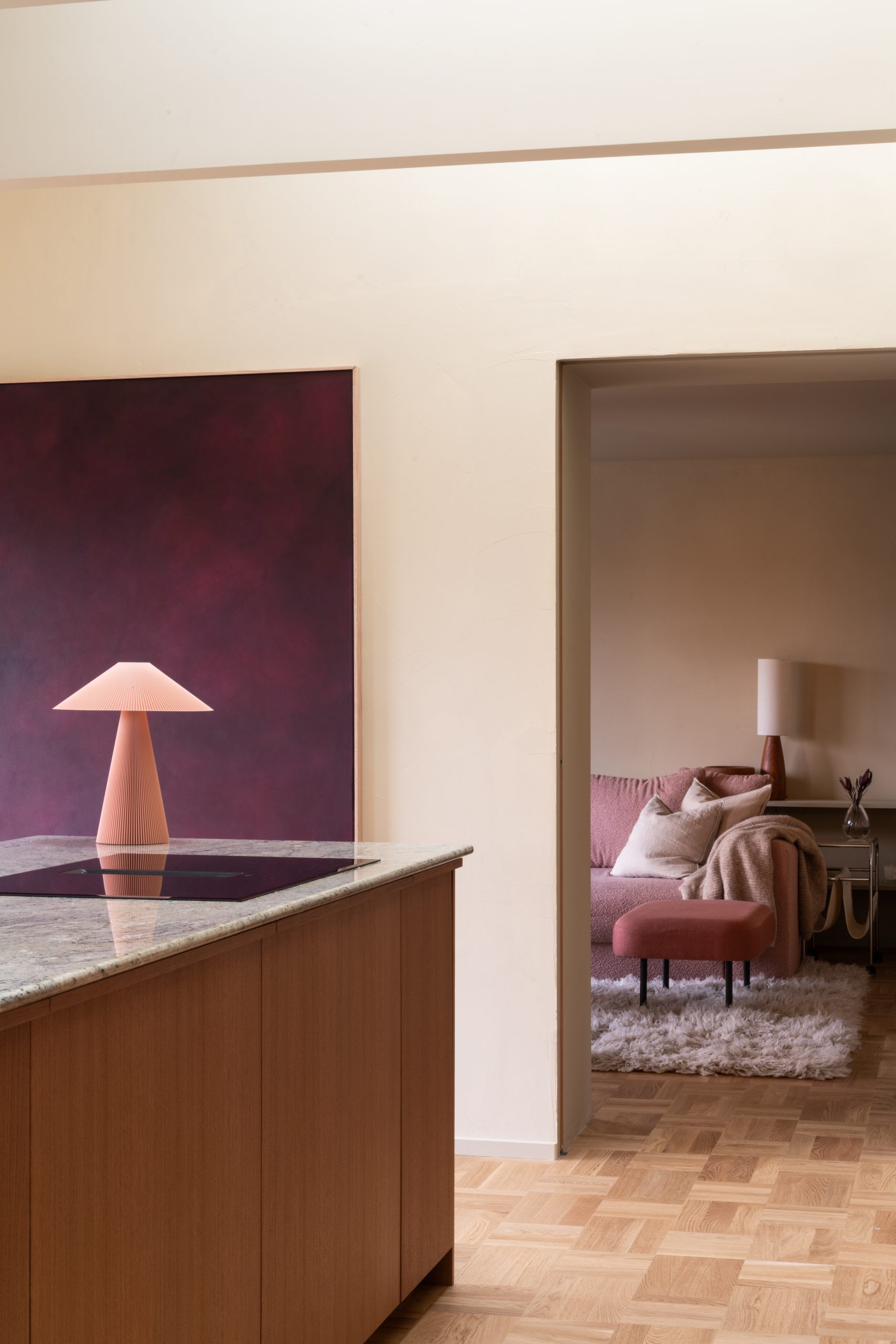 Moderne interiør med åpen kjøkkenløsning og stue. Et marmorbelagt kjøkkenøy med trepanel står i forgrunnen, mens en rød moderne lampe og kunstverk vises mot veggen. I bakgrunnen er en koselig stue med rosa sofa og teppe synlig gjennom en døråpning