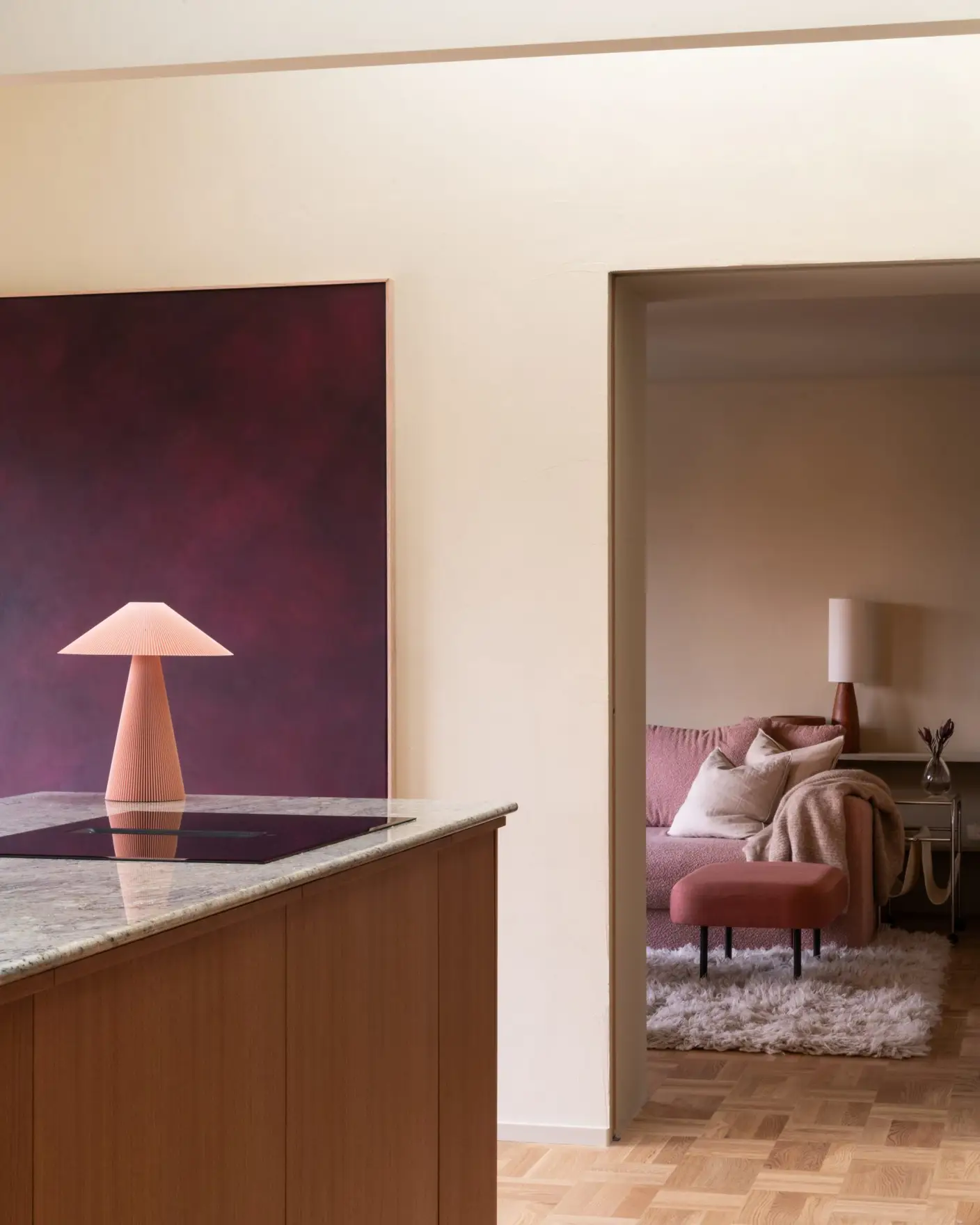 Moderne interiør med åpen kjøkkenløsning og stue. Et marmorbelagt kjøkkenøy med trepanel står i forgrunnen, mens en rød moderne lampe og kunstverk vises mot veggen. I bakgrunnen er en koselig stue med rosa sofa og teppe synlig gjennom en døråpning