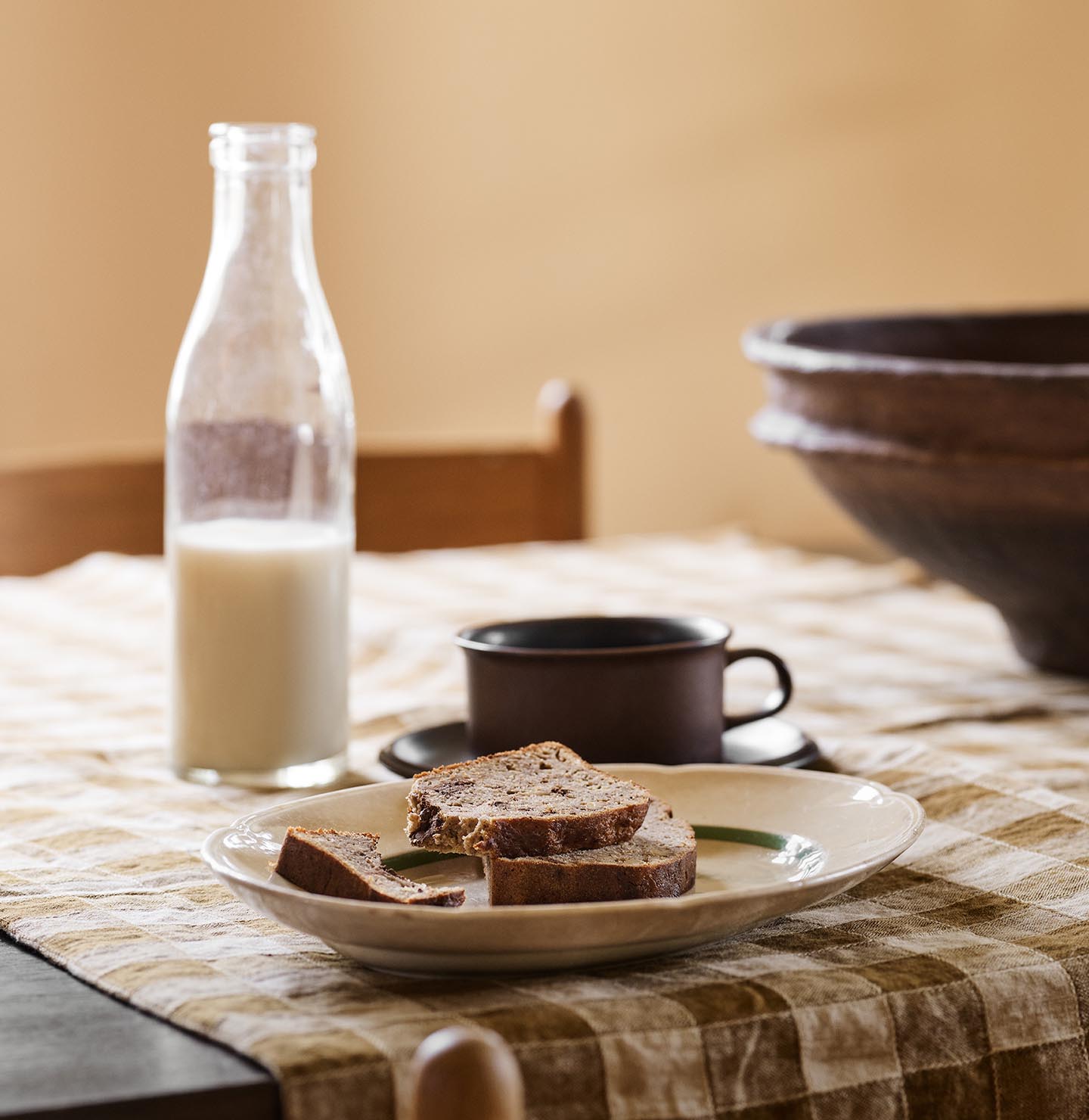 Table with bread, a cup, and a milk bottle on a checkered cloth against a ochre yellow background painted in with the Jotun colour 11202 Mild Ochre.
