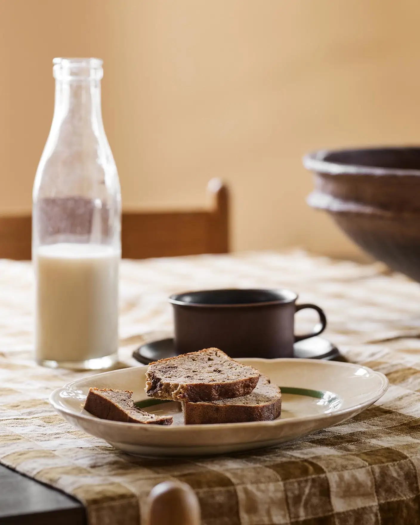 Table with bread, a cup, and a milk bottle on a checkered cloth against a ochre yellow background painted in with the Jotun colour 11202 Mild Ochre.