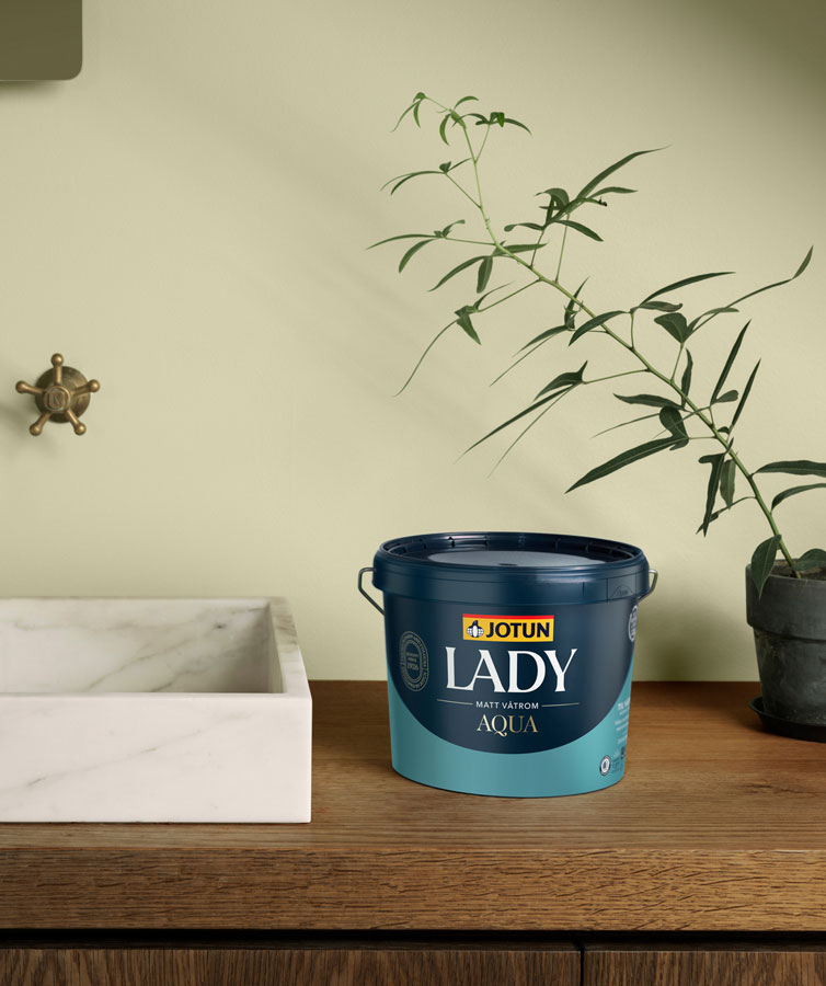 Malingbøtte merket "Jotun Lady Aqua" på en trebenk ved siden av en hvit marmorvask og potteplante i et minimalistisk miljø