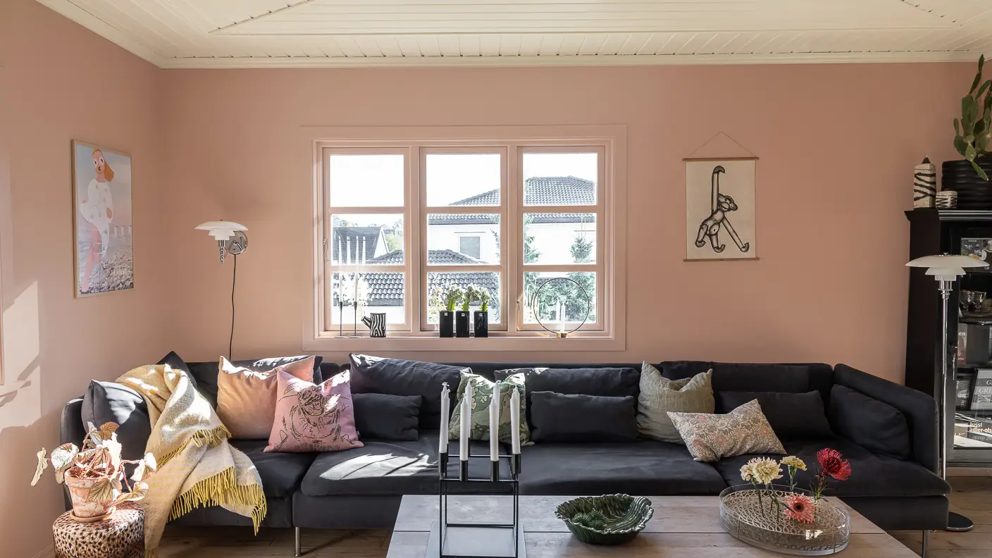 Vardagsrum med mörkgrå soffa med kuddar i ljusa toner, rosa väggar och fönsterkarmar målade i Jotun 20362 Pink Ambience, samt tak i beige Jotun 12313 Soft Dunes.