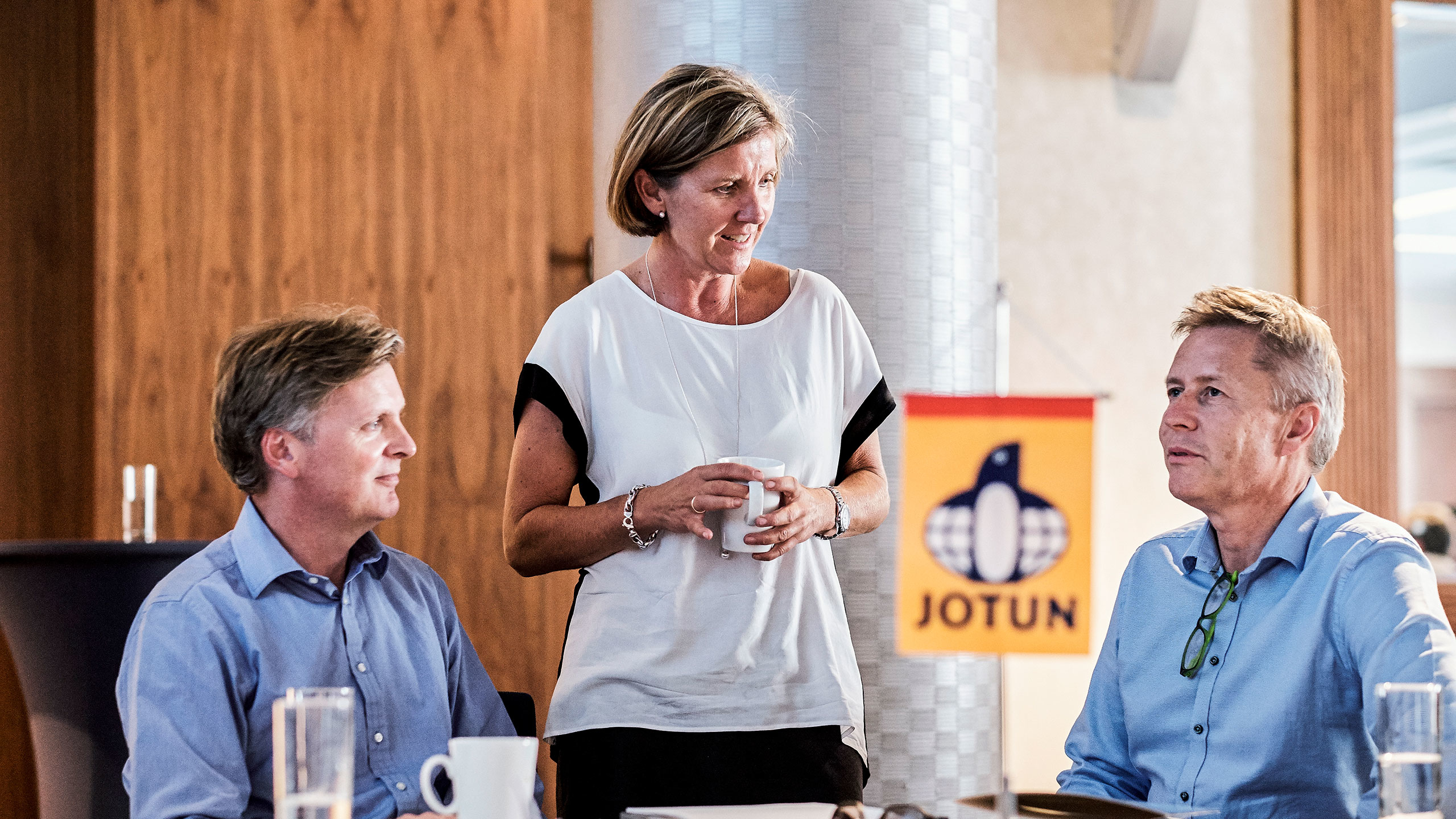 Tre personer diskuterar vid ett bord på ett kontor, med en Jotun-flagga synlig i förgrunden