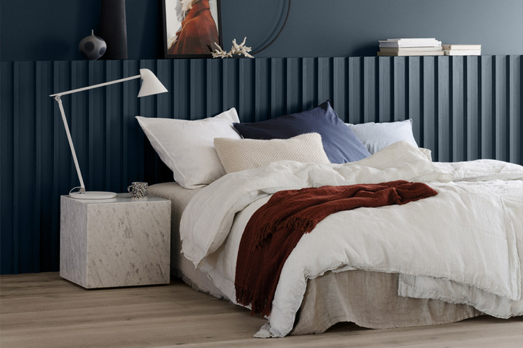 Moderne soverom med en luksuriøs seng, lagdelte hvite og marineblå puter, et varmt rødt pledd, og et elegant marmor-sidebord med en moderne hvit lampe, satt mot en mørkeblå panelvegg