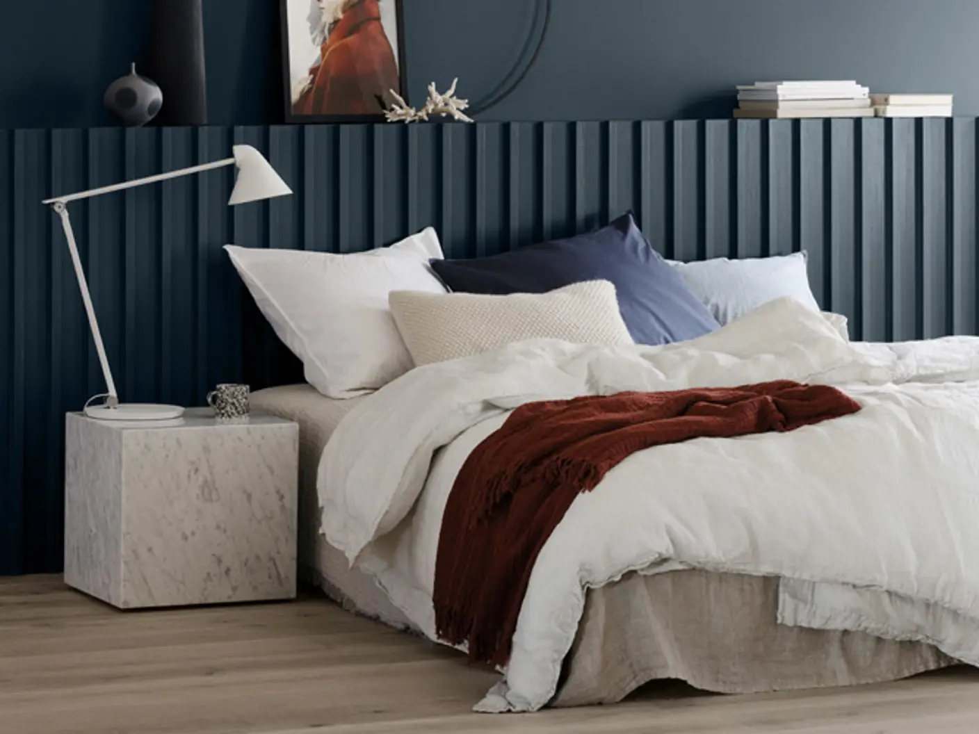 Moderne soverom med en luksuriøs seng, lagdelte hvite og marineblå puter, et varmt rødt pledd, og et elegant marmor-sidebord med en moderne hvit lampe, satt mot en mørkeblå panelvegg