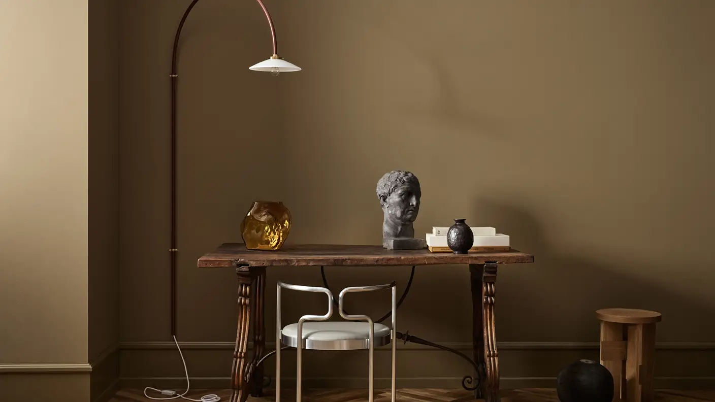 Skrivebord i mørkt treverk med stol i blankt stål, byste på bordplate og Valerie Objects lampe på vegg malt i gyllenbrune Jotun 1974 Golden Walnut.