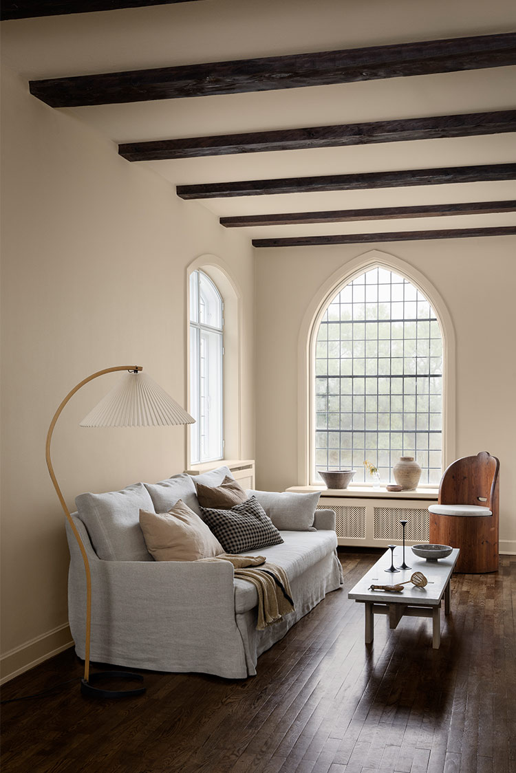 Koselig stue med en lys grå sofa, forskjellige puter, og en gulvlampe i tre. Et minimalistisk sofabord har dekorative gjenstander, plassert på mørke tregulv. Et stort buet vindu med rutemønster slipper inn naturlig lys på beige vegger malt i Jotun 1303 Observe