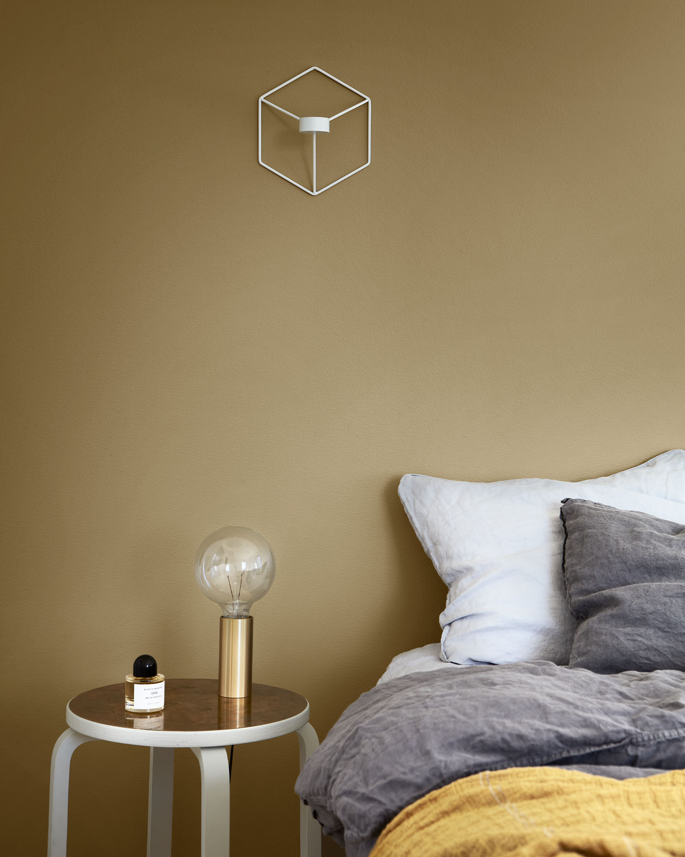 Mysig sovrumshörna med ett grått och senapsgult bäddset, ett litet runt sidobord med en guldlampa och en parfymflaska, mot en dämpad gulgrön vägg målad med Jotun 1864 Anonymous.