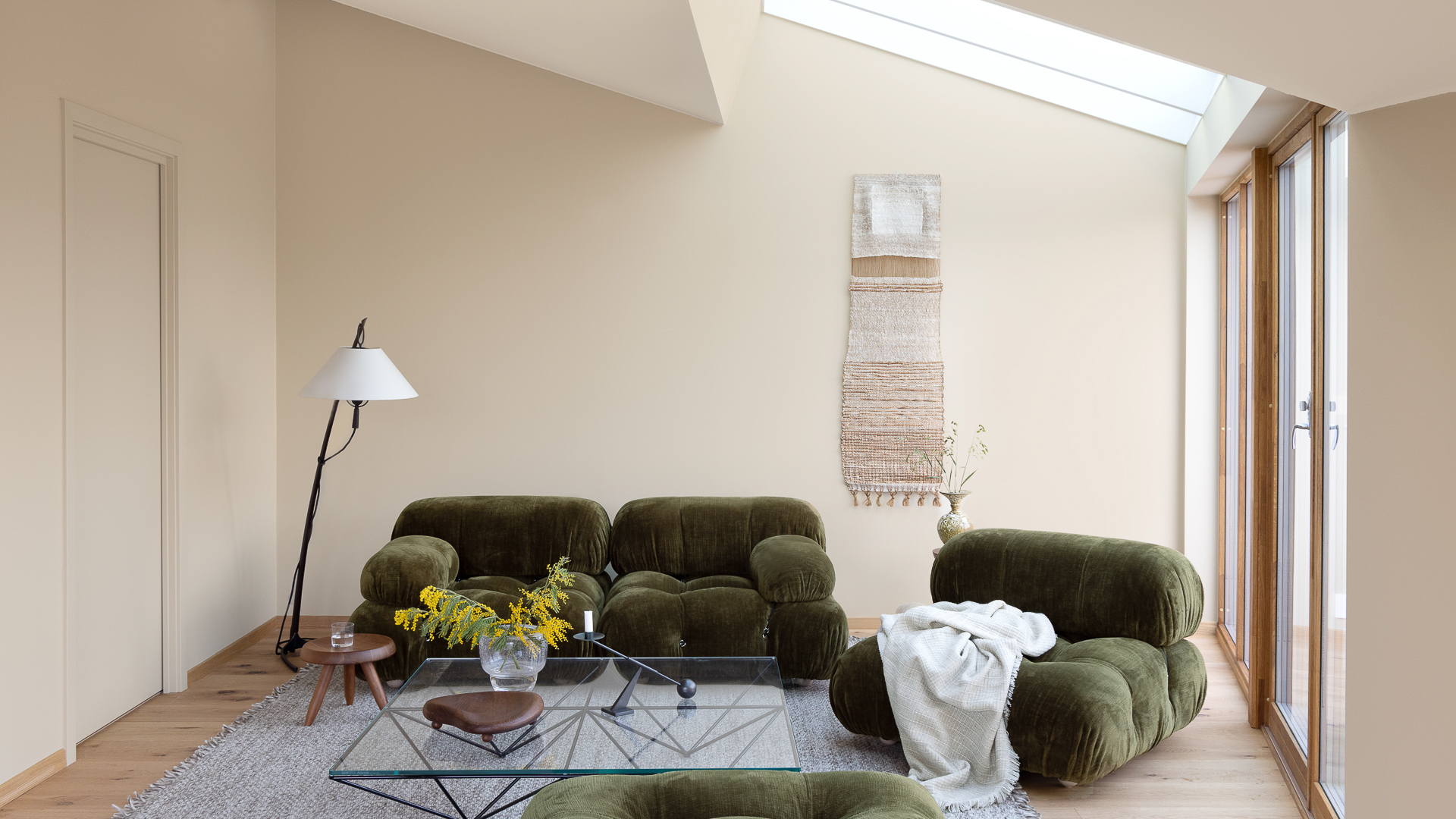 Beige stue malt i JOTUN 1303 Observe og JOTUN 12313 Soft Dunes, møblert med grønn sofa og stol, moderne glassbord, en stålampe og vevarbeid i naturtoner på veggen.