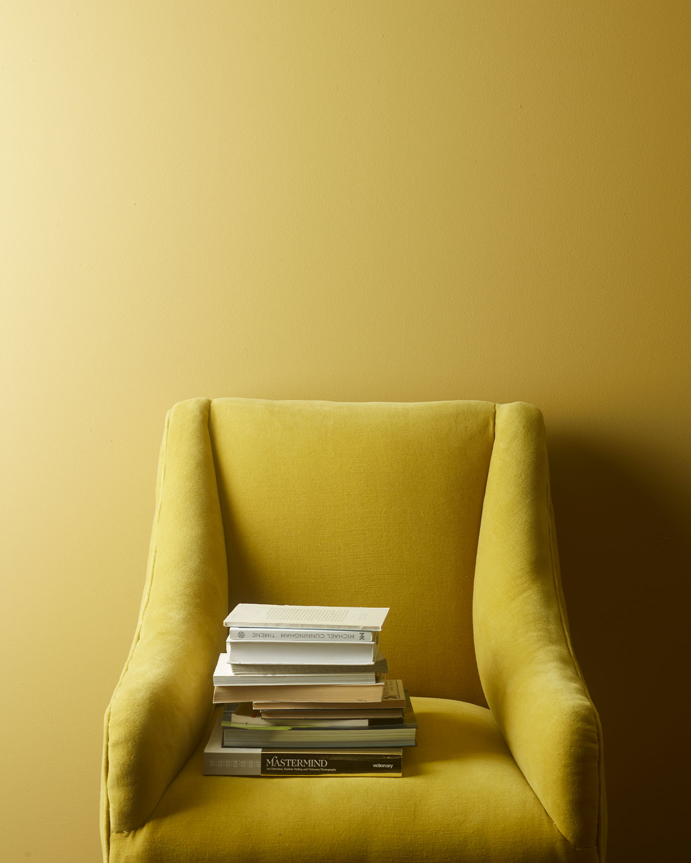 Mustard yellow armchair with a stack of books resting on the seat, set against a muted, greenish yellow wall painted with Jotun 0288 Mexico.